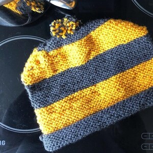 Può includere: Un sottopentola lavorato a maglia a strisce gialle e grigie con un pompon in cima.