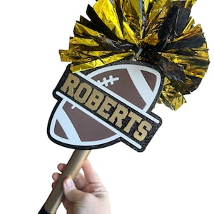 May include: A gold and black pom-pom attached to a football-shaped sign with the name "ROBERTS" in gold lettering. The sign is brown and white with a gold handle. The pom-pom is gold and black.