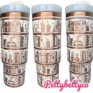 May include: A white and copper travel mug with a white lid and handle. The mug features a design of books on shelves with a copper finish. The text "Pettybettyco" is printed on the bottom of the mug.