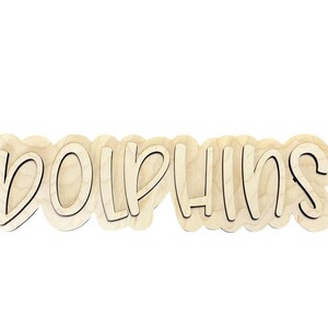 May include: Wooden sign with the word "DOLPHINS" in a playful, layered font. The letters are light-colored wood with a dark outline, set against a white background. The sign has a wavy, cloud-like shape.