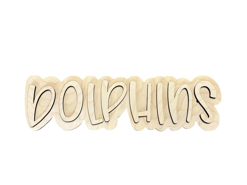 May include: Wooden sign with the word "DOLPHINS" in a playful, layered font. The letters are light-colored wood with a dark outline, set against a white background. The sign has a wavy, cloud-like shape.