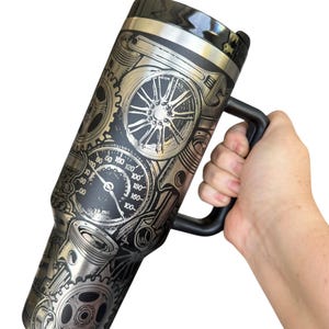 Może przedstawiać: Czarny kubek termiczny ze stali nierdzewnej z srebrnym wzorem części samochodowych, w tym prędkościomierza, kół zębatych i tłoków. Kubek ma czarny uchwyt.