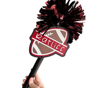 May include: A handheld spirit stick featuring a dark maroon metallic pom-pom attached to a black handle. A flat, shield-shaped sign is mounted below the pom-pom, displaying a brown football graphic and the name Ratliff in white script lettering.