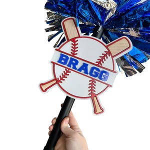May include: A baseball-themed spirit stick with a white baseball design, red stitching, and crossed bats. The word "BRAGG" is displayed in blue. The stick has a black handle and is topped with blue and silver pom-poms.