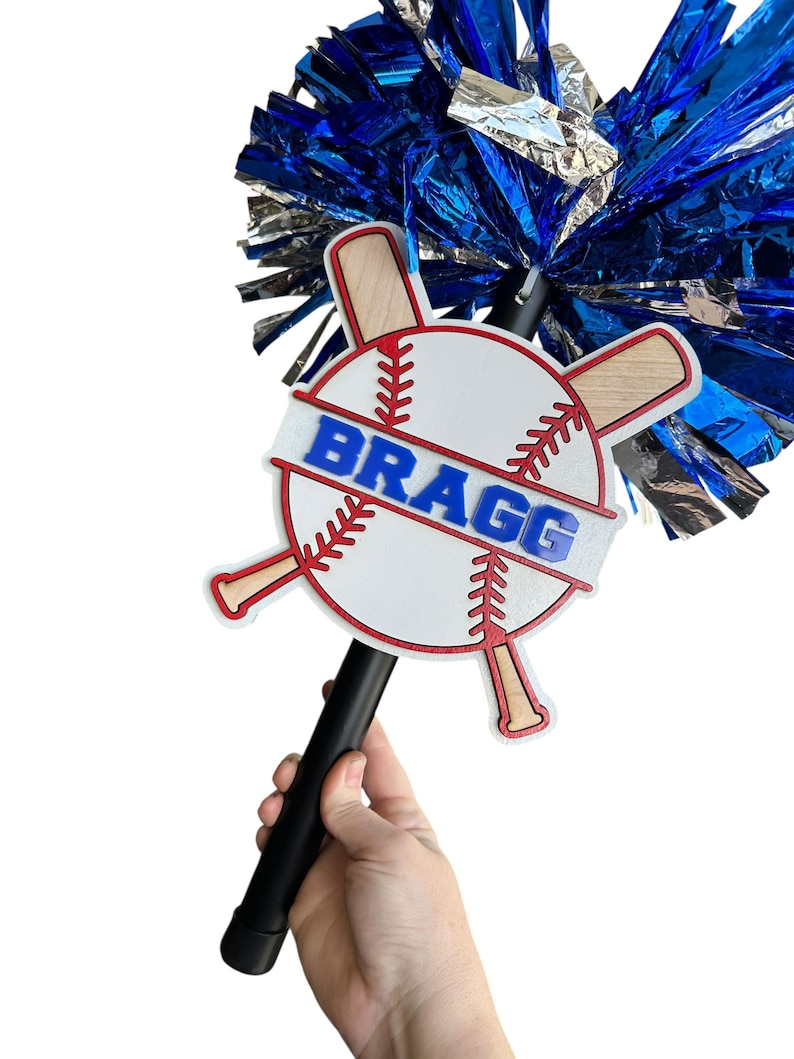 May include: A baseball-themed spirit stick with a white baseball design, red stitching, and crossed bats. The word "BRAGG" is displayed in blue. The stick has a black handle and is topped with blue and silver pom-poms.