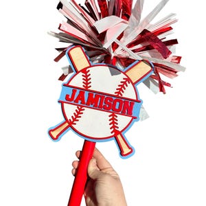May include: A red and white baseball-themed spirit stick with the name "JAMISON" in blue. The stick features a baseball design with crossed bats and a red handle. The top has red, white, and silver streamers.