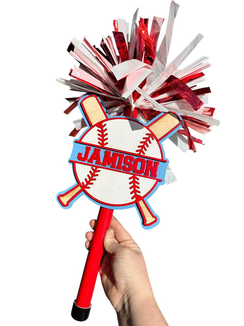 May include: A red and white baseball-themed spirit stick with the name "JAMISON" in blue. The stick features a baseball design with crossed bats and a red handle. The top has red, white, and silver streamers.