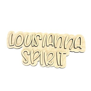 May include: A wooden sign with the words "LOUISIANA SPIRIT" cut out in a cursive font. The sign is made of light-colored wood with a dark outline, and the edges have a scalloped design.