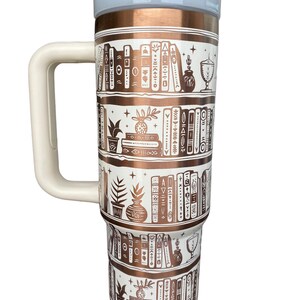 May include: A white and copper travel mug with a white handle. The mug features a design of books on shelves with plants and other decorative elements. The mug has a copper lid with a white top.