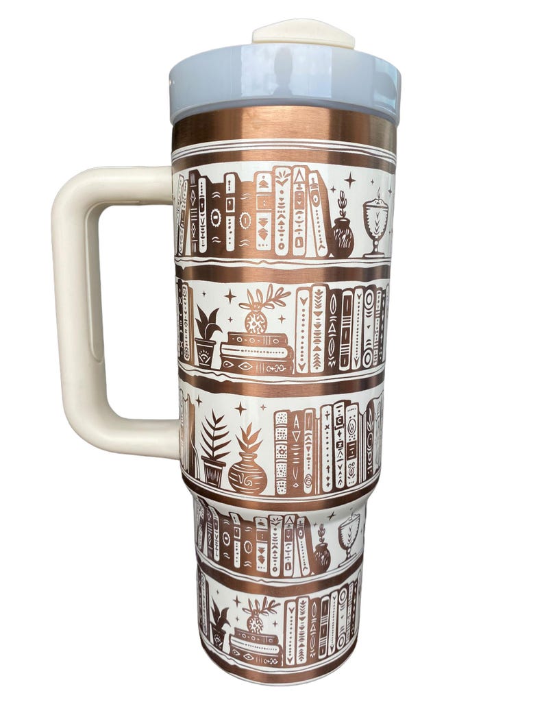 May include: A white and copper travel mug with a white handle. The mug features a design of books on shelves with plants and other decorative elements. The mug has a copper lid with a white top.