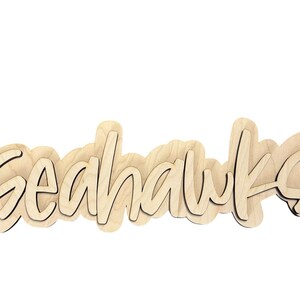 May include: Wooden sign with the word "Seahawks" in a cursive font. The sign is made of light-colored wood with a dark outline, and the letters are layered for a 3D effect. The sign is a decorative item.