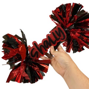 May include: A pair of cheerleading pom-poms featuring metallic red and black plastic streamers. A central handle displays the name Shally in black script lettering set against a red glittered background, creating a bold, high-contrast design.