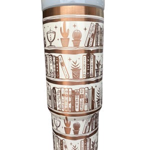 May include: A white and copper tumbler with a bookshelf design. The tumbler features a pattern of books, plants, and stars in a copper color on a white background.