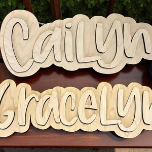May include: Two wooden name signs, one reading "Cailyn" and the other "GraceLyn". The names are in a layered, cursive font, with a light wood finish. The signs are on a dark brown surface, with greenery and flowers in the background.