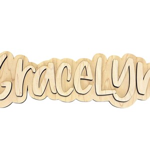 May include: Wooden name cutout spelling "Gracelyn" in a layered, bubbly font. The letters are light brown with a darker outline, creating a 3D effect. The cutout is designed to be a decorative item, perfect for a nursery or child's room.