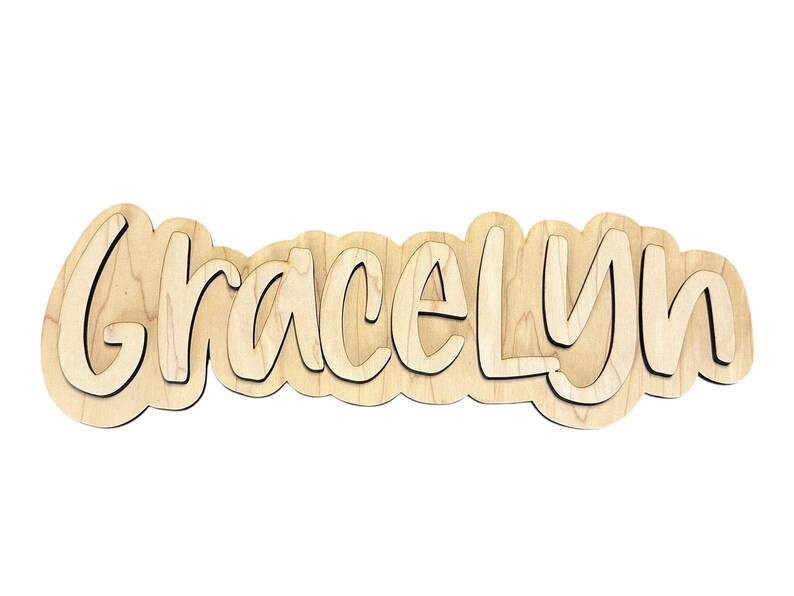 May include: Wooden name cutout spelling "Gracelyn" in a layered, bubbly font. The letters are light brown with a darker outline, creating a 3D effect. The cutout is designed to be a decorative item, perfect for a nursery or child's room.