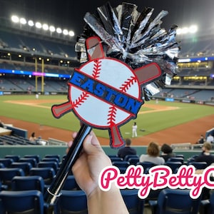 Puede incluir: Un abanico con temática de béisbol con un mango negro, un diseño de béisbol y la palabra "EASTON" en azul. El abanico incluye un pompón plateado y negro y se sostiene frente a un estadio de béisbol.