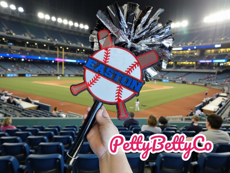 May include: A baseball-themed fan with a black handle, a baseball design, and the word "EASTON" in blue. The fan includes a silver and black pom-pom and is held in front of a baseball stadium.