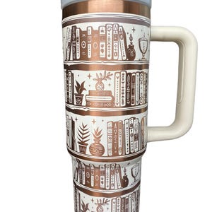 May include: A white and copper travel mug with a white handle. The mug has a design of bookshelves with books, plants, and other decorative items.
