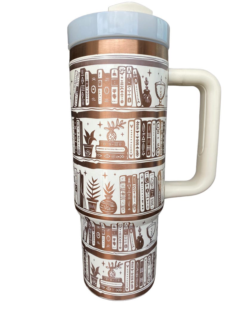 May include: A white and copper travel mug with a white handle. The mug has a design of bookshelves with books, plants, and other decorative items.