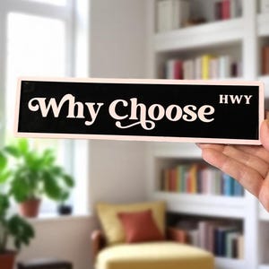 May include: A black and pink sign with the words "Why Choose" in white script. The word "HWY" is in white block letters. The sign is being held up in front of a blurred background of a room with a window and bookshelves.