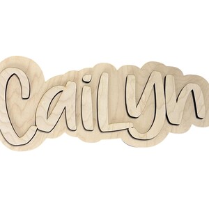 May include: Wooden name cutout spelling "Cailyn" in a playful font. The name is layered, with a light wood color and a dark outline, set against a white background. Ideal for nursery decor.