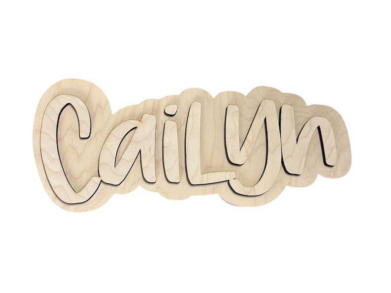 May include: Wooden name cutout spelling "Cailyn" in a playful font. The name is layered, with a light wood color and a dark outline, set against a white background. Ideal for nursery decor.