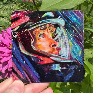 May include: A square sticker featuring a vibrant, glittery illustration of an astronaut looking upward. The artwork displays a person with orange-toned skin inside a dark helmet, surrounded by swirling streaks of blue, purple, and pink cosmic light.