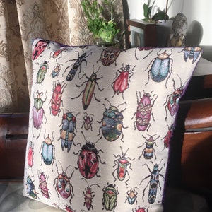 Op de afbeelding: Een wit kussen met een paarse rand heeft een herhalend patroon van kleurrijke kevers en insecten. Het kussen is gemaakt van een zachte, textuurrijke stof.