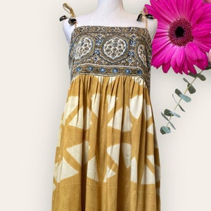 May include: A mustard yellow sleeveless dress with a white geometric pattern and a blue and brown floral print border. The dress has a gathered waist and a tie at the shoulders.