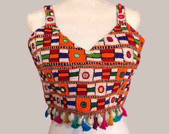 May include: An orange, white, red, green, and blue embroidered crop top with a V-neck and shoulder straps. The top features a geometric pattern with small mirrors and colorful tassels along the bottom edge. The garment is displayed on a mannequin.
