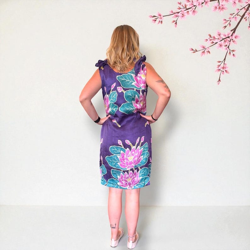 May include: A sleeveless purple dress with a floral lotus print in pink and teal. The dress has a knee-length hemline and shoulder ties. The model is standing with their hands on their hips.