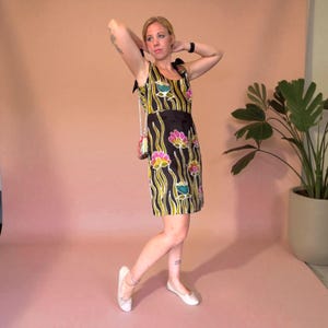May include: A sleeveless black dress with a floral pattern in pink, yellow, and teal. The dress has a black ribbon belt and black shoulder straps. A small, patterned purse hangs from the shoulder. The dress is knee-length.
