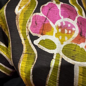 May include: Close-up of a black fabric with a floral pattern. The design features large pink flowers with yellow and orange centers, and green and yellow leaf-like shapes. White outlines and accents add detail.