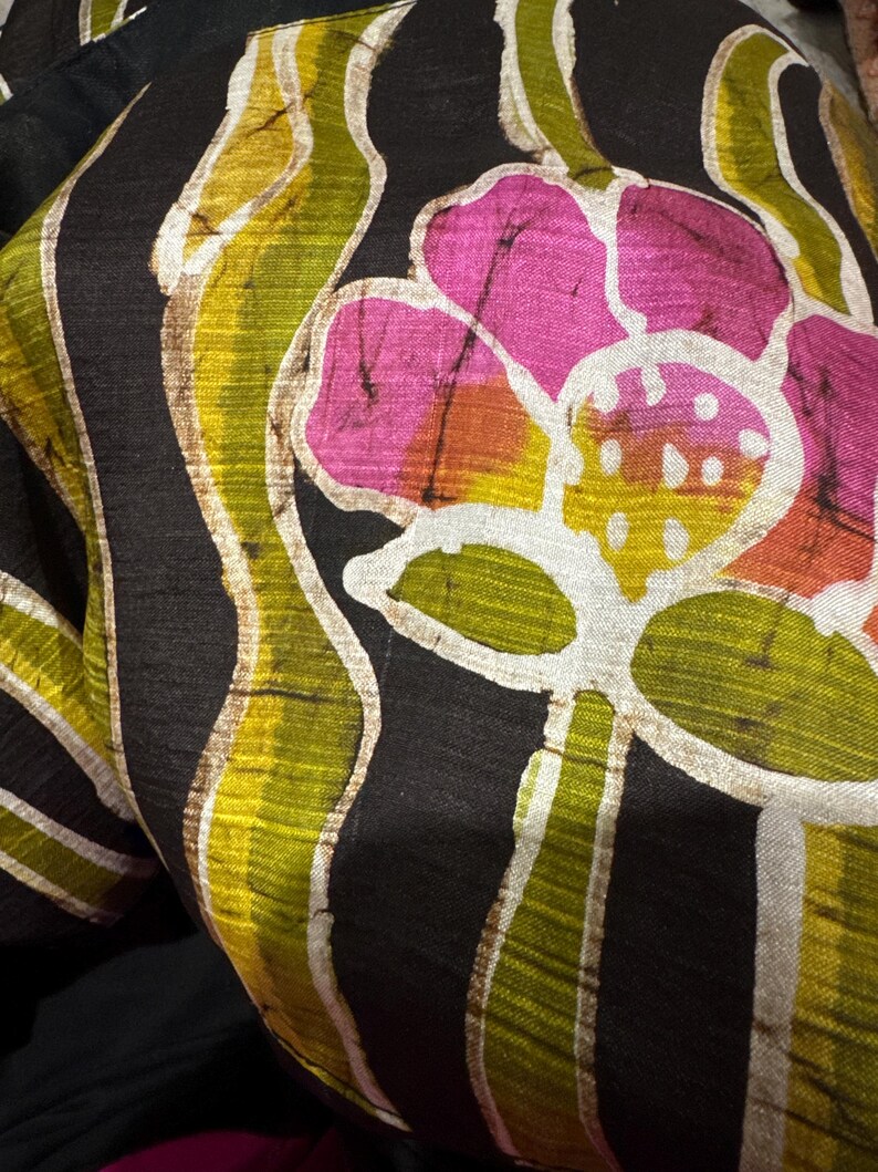 May include: Close-up of a black fabric with a floral pattern. The design features large pink flowers with yellow and orange centers, and green and yellow leaf-like shapes. White outlines and accents add detail.