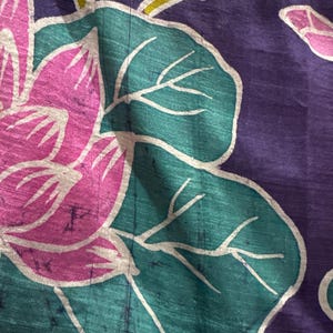 May include: Close-up of a textile with a floral pattern. The design features a large pink flower with white outlines, teal leaves, and purple background. The fabric has a textured appearance.