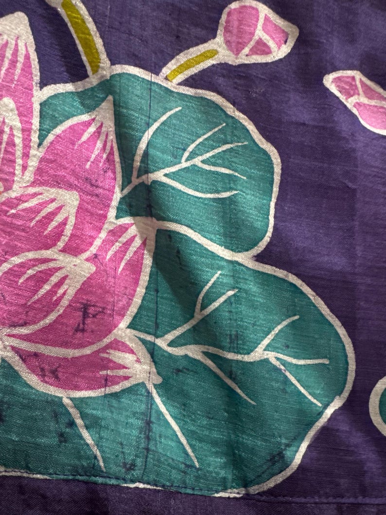 May include: Close-up of a textile with a floral pattern. The design features a large pink flower with white outlines, teal leaves, and purple background. The fabric has a textured appearance.
