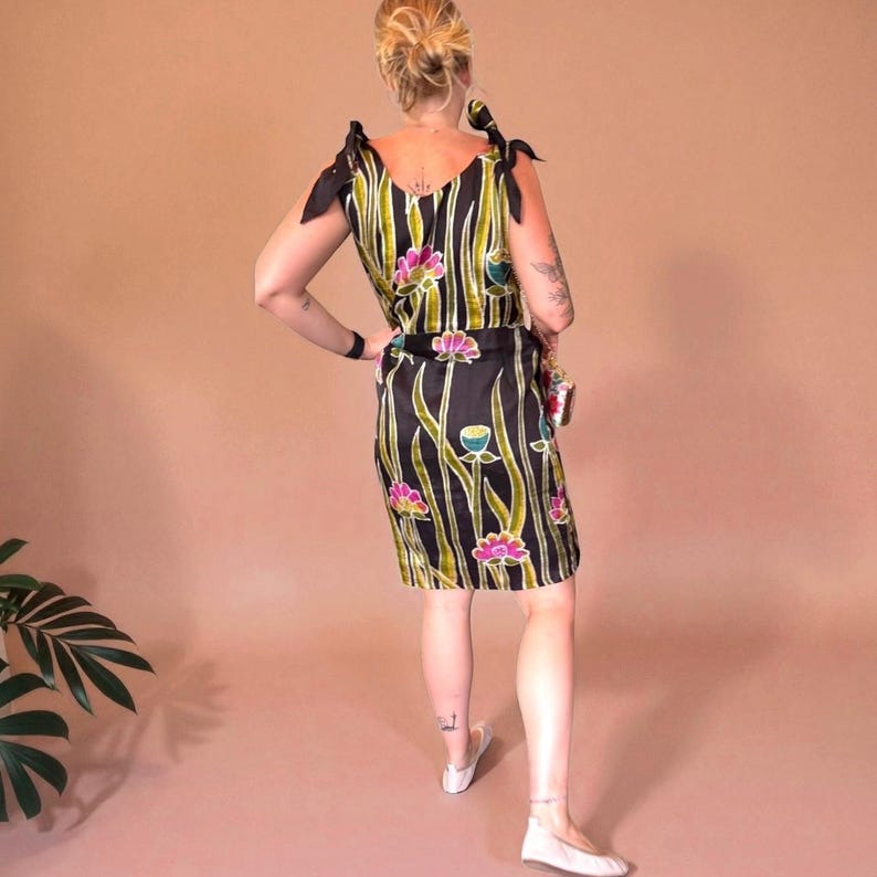 May include: A black dress with a floral pattern. The dress features a V-neckline, tie straps, and a fitted silhouette. The floral design includes pink, green, and blue accents. The dress is knee-length and has a matching belt.