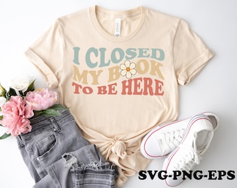 May include: A beige t-shirt with the text "I closed my book to be here" in a retro font with a daisy graphic. The shirt is laid out on a white surface with a pair of white sneakers and a pair of grey jeans.