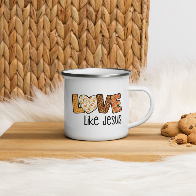 May include: White enamel mug with a silver rim and handle. The mug features a patchwork design of the word "LOVE" with a heart in the "O". The text "Like Jesus" is printed below the word "LOVE".