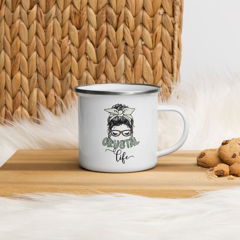Puede incluir: Taza de esmalte blanco con una ilustraci&oacute;n en blanco y negro de una mujer con un mo&ntilde;o y gafas. El texto "Crystal Life" est&aacute; escrito en verde debajo de la ilustraci&oacute;n.