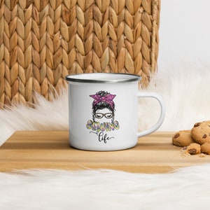 Pu&ograve; includere: Tazza in smalto bianco con bordo e manico neri. La tazza presenta un'illustrazione colorata di una donna con un chignon e occhiali, con il testo "Sewing Life" sotto.