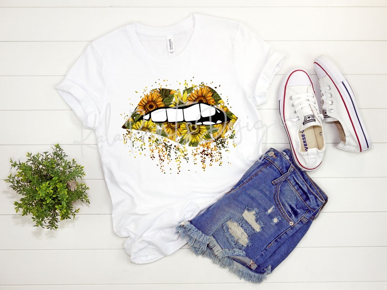 May include: White t-shirt with a graphic of a pair of lips covered in sunflowers. The lips are slightly open, showing teeth. The background is a white wood surface.