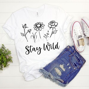 May include: White t-shirt with a black floral design and the text "Stay Wild". The shirt is laid out on a white wooden surface with a pair of white sneakers and denim shorts.