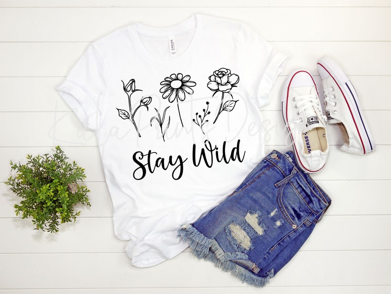 May include: White t-shirt with a black floral design and the text "Stay Wild". The shirt is laid out on a white wooden surface with a pair of white sneakers and denim shorts.