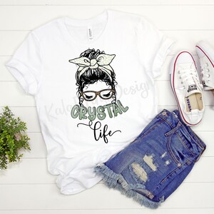Puede incluir: Camiseta blanca con un gr&aacute;fico de una mujer con un mo&ntilde;o desordenado, gafas y una diadema. El texto "CRYSTAL life" est&aacute; debajo del gr&aacute;fico.