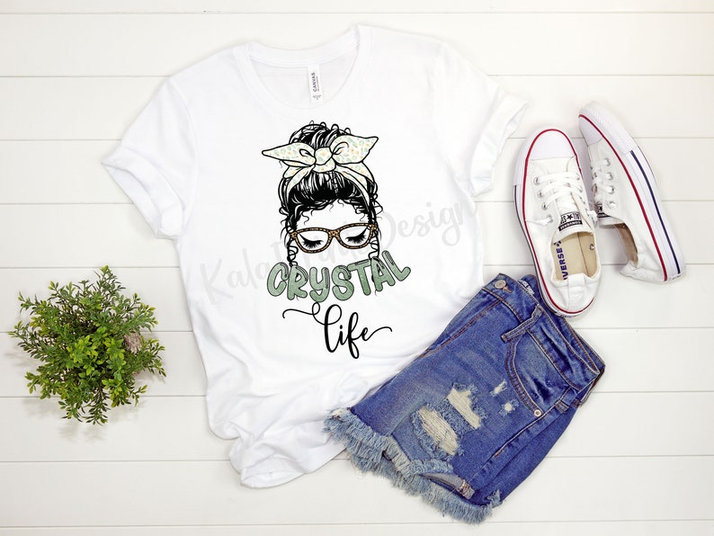 Puede incluir: Camiseta blanca con un gr&aacute;fico de una mujer con un mo&ntilde;o desordenado, gafas y una diadema. El texto "CRYSTAL life" est&aacute; debajo del gr&aacute;fico.