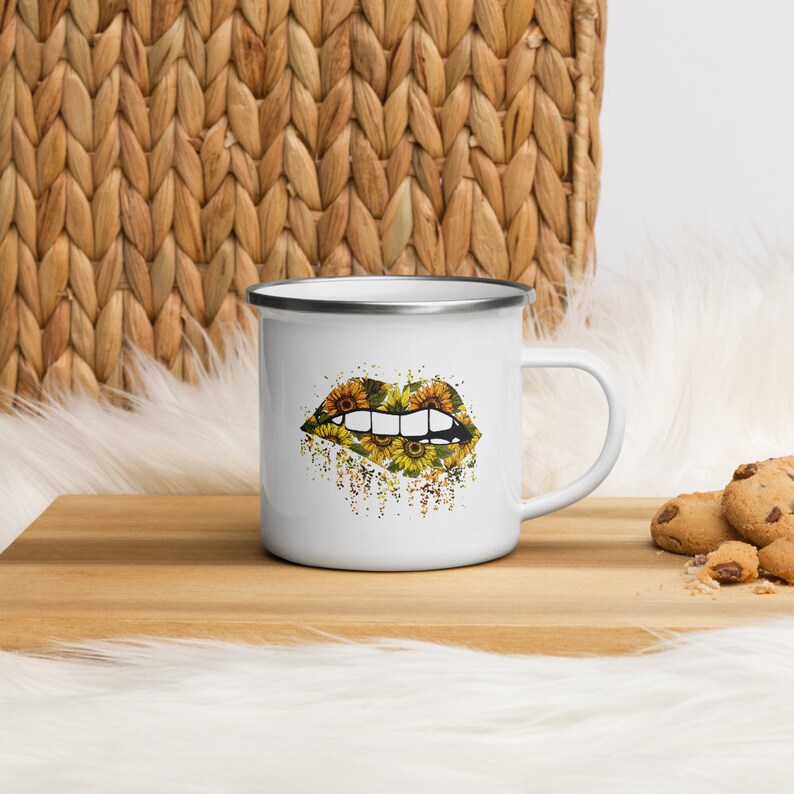 May include: White enamel mug with a black outline. The mug features a graphic of a pair of lips with a sunflower pattern.