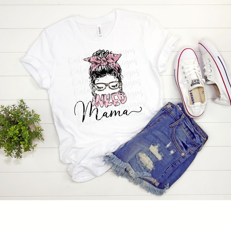 May include: White t-shirt with a graphic of a woman with a bun, glasses, and a pink leopard print bandana. The text "INKED MAMA" is written below the graphic.