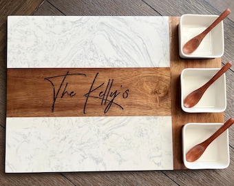 Puede incluir: Una bandeja de servir de mármol blanco y madera con tres cuencos pequeños blancos y cucharas de madera. La parte de madera de la bandeja tiene el texto "The Kelly's" grabado.